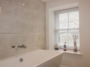 A bathroom with a white bathtub under a window with blinds and decorative items on the windowsill at The Crows Nest in Mevagissey