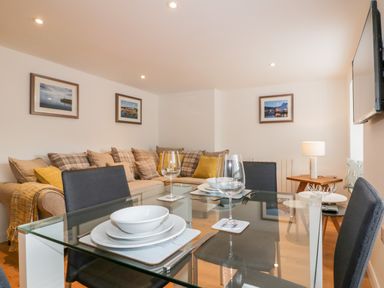 A dining area with a glass table set with plates and glasses and a living space with a sofa and framed pictures on the wall at The Crows Nest in Mevagissey