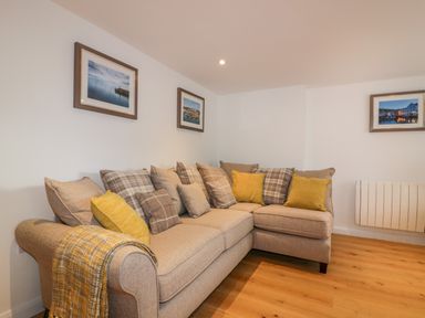 A beige sectional sofa with yellow and plaid cushions and a throw in a room with wooden floor and framed pictures on the wall at The Crows Nest in Mevagissey