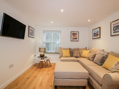 A living room with a beige sectional sofa and ottoman a wall-mounted tv and framed pictures at Captains Cabin in Mevagissey