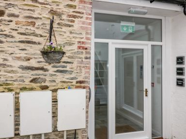 A stone wall with a hanging basket and utility boxes next to a glass door with emergency exit signs at Captains Cabin in Mevagissey