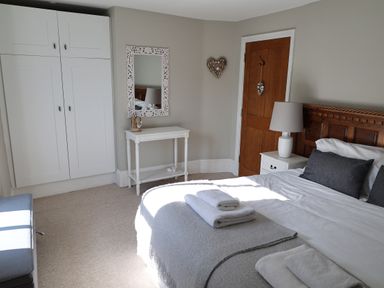 A bedroom with a wooden bed, white bedding, gray pillows, a white nightstand with a lamp, a white table with a mirror above, a wardrobe, and a wooden door at The Penthouse Lynmouth Bay in Lynton