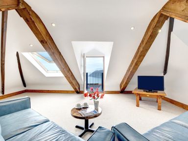 An attic living room with wooden beams a small table with flowers and cups two blue sofas a television on a wooden stand and a door opening to a balcony at The Penthouse Lynmouth Bay in Lynton