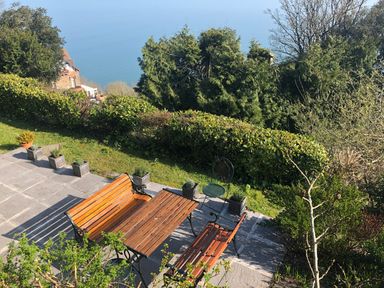 An outdoor patio with wooden benches a table metal chairs and potted plants overlooking trees and a distant sea view at The Penthouse Lynmouth Bay in Lynton