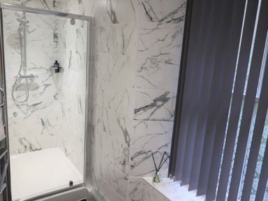 A bathroom with a glass shower enclosure and marble tiled walls next to a window with vertical blinds at The Penthouse Lynmouth Bay in Lynton