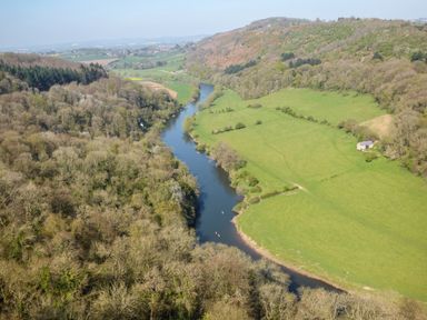 A river flowing through a valley with wooded hills on one side and green fields with a small building on the other at The Lookout in Llangrove