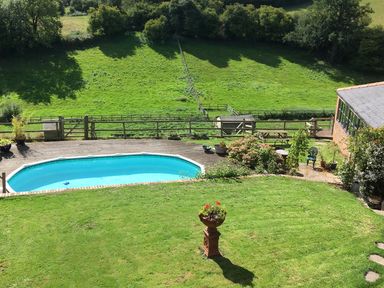 A backyard with a swimming pool surrounded by wooden decking a lawn with a flower pot a garden area with plants a picnic table and a brick building at The Lookout in Llangrove