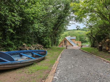 A gravel driveway leading to a wooden house with a blue boat and stacked logs on the sides at The Lookout in Llangrove