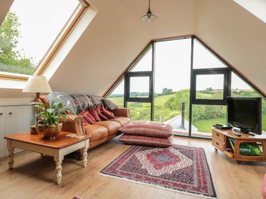 A living room with a leather sofa red pillows patterned rug and a television near a large triangular window at The Lookout in Llangrove