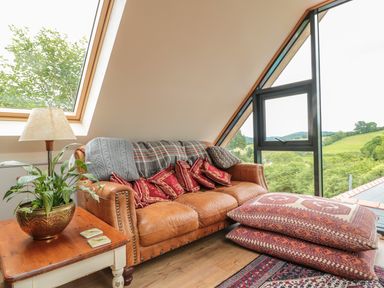 A living area with a leather sofa, patterned cushions, a wooden side table with a lamp and plant, and large windows overlooking green hills at The Lookout in Llangrove