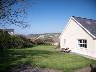 A garden with green grass bushes and a wooden picnic table next to a house with hills and houses in the background at Bramble Cottage in Newgale