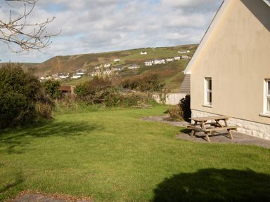 A backyard with green grass a wooden picnic table and a beige house with hills and houses in the background at Bramble Cottage in Newgale