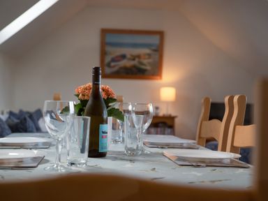 A dining table set with plates glasses a wine bottle and a flower pot in a room with wooden chairs and a painting on the wall at Bramble Cottage in Newgale