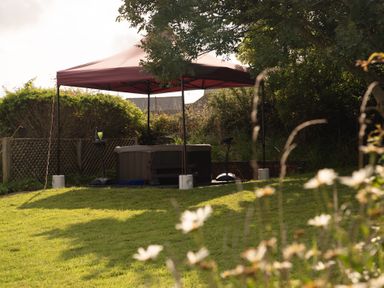 An outdoor area with a hot tub under a canopy surrounded by grass and trees at Bramble Cottage in Newgale