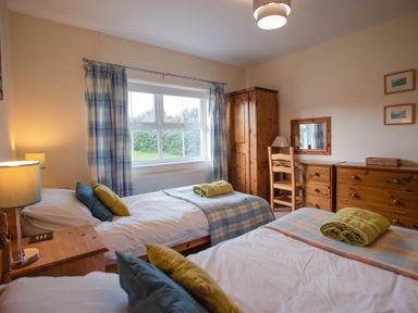 A bedroom with two single beds a wooden wardrobe a dresser and a window with plaid curtains at Bramble Cottage in Newgale