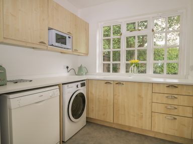 A kitchen with wooden cabinets a white washing machine dishwasher microwave toaster kettle and a window with yellow flowers on the countertop at Thames Reach in Wallingford
