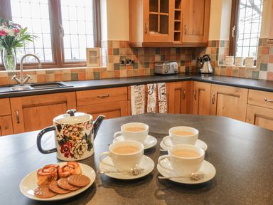 A kitchen with wooden cabinets a teapot four cups of tea and a plate of biscuits and pastries on a countertop at Ty Berllan in Caersws