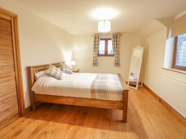 A bedroom with a wooden bed with pillows and a blanket beside a window with curtains and a standing mirror at Ty Berllan in Caersws