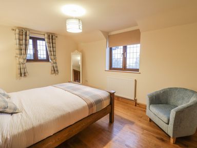 A bedroom with a wooden bed with white and plaid bedding a blue armchair wooden floor and two windows with curtains at Ty Berllan in Caersws