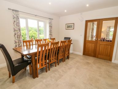 A dining room with a wooden table surrounded by ten chairs and floral curtains at Ochr Y Garth in Maenan