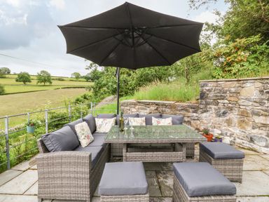 An outdoor seating area with a glass table a large umbrella and cushioned wicker furniture on a stone patio at Ochr Y Garth in Maenan