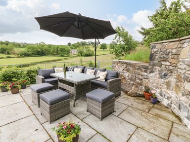 An outdoor patio with rattan furniture seating a table under a large umbrella with a stone wall and countryside view at Ochr Y Garth in Maenan