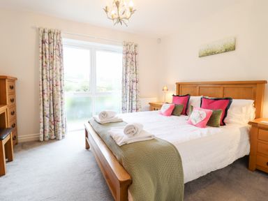 A bedroom with a wooden bed frame white bedding and colorful pillows next to wooden bedside tables and floral curtains at Ochr Y Garth in Maenan