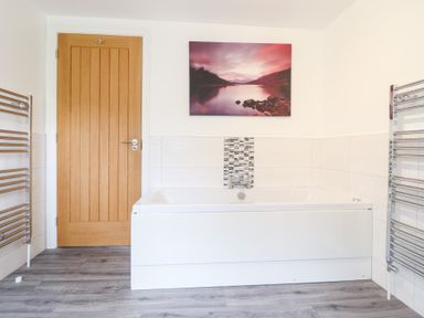 A bathroom with a white bathtub wooden door two heated towel rails and a landscape picture above the bathtub at Ochr Y Garth in Maenan