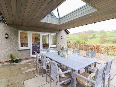 An outdoor covered patio with a dining table and chairs overlooking a green landscape at Ochr Y Garth in Maenan