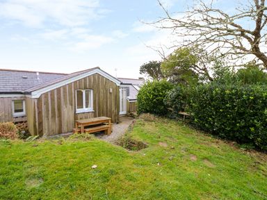 A wooden exterior of a house with a bench table set on a gravel path and green grass garden with bushes and a tree at Oreos Cottage in St Agnes