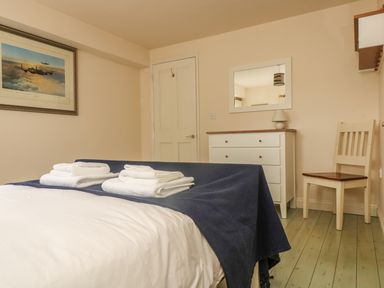 A bedroom with a bed covered by white sheets and a blue blanket two stacks of towels on the bed a white dresser with a lamp a mirror above the dresser a wooden chair and a door on the beige walls at Oreo's Cottage in St Agnes