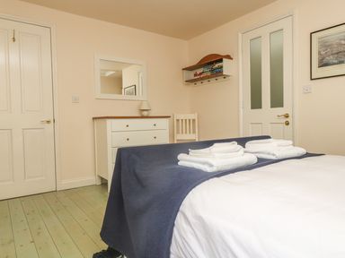 A bedroom with a bed covered with a blue blanket and white towels on top a white dresser with a lamp a wooden chair a wall shelf with books two closed doors and a framed picture at Oreos Cottage in St Agnes