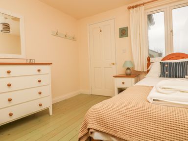 A bedroom with a bed, a nightstand with a lamp, a chest of drawers with a mirror, and a door at Oreos Cottage in St Agnes