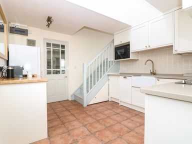 A kitchen with white cabinets a wooden countertop a white farmhouse sink a microwave under the stairs and a tiled floor at Oreos Cottage in St Agnes