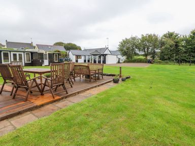 An outdoor area with wooden tables and chairs on a wooden deck next to a large grassy lawn with white and brown buildings in the background at The Lodge in Rosemarket