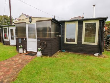 A dark wooden building with two windows and a white door surrounded by grass at The Lodge in Rosemarket