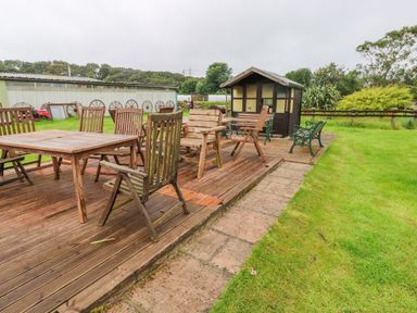 An outdoor wooden deck with multiple wooden chairs and tables near a small shed on grass at The Lodge in Rosemarket