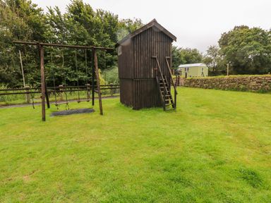 A wooden playhouse with stairs and a swing set on grass near a stone wall and trees at The Lodge in Rosemarket