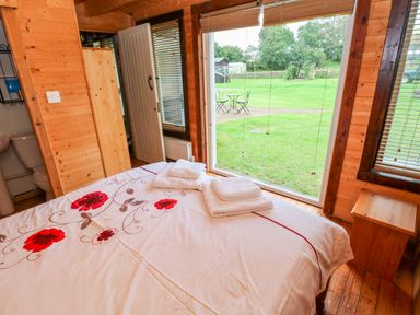 A bedroom with a bed covered in white bedding with red flowers folded towels on top a wooden nightstand and a large window showing a green lawn with outdoor furniture at The Lodge in Rosemarket