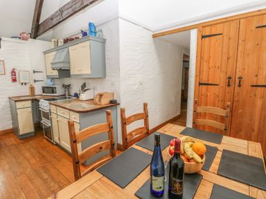 A kitchen with wooden dining table and chairs a bowl of fruit and two bottles on the table and cabinets with appliances in a cottage at Heather Cottage Rosemarket