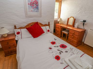 A bedroom with a bed with floral bedding beside a wooden nightstand and a wooden dresser with a mirror at Heather Cottage in Rosemarket