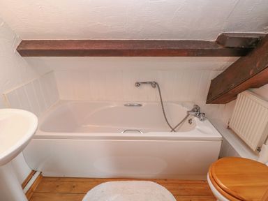 A bathroom with a bathtub under a wooden beam a white sink a toilet with a wooden seat and a radiator at Heather Cottage in Rosemarket