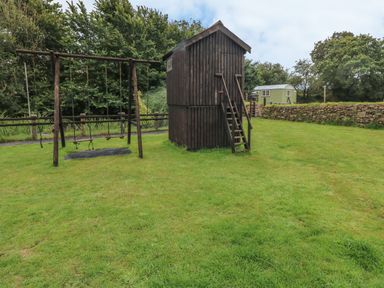 A backyard with a wooden swing set and a small wooden playhouse on grass at Heather Cottage in Rosemarket