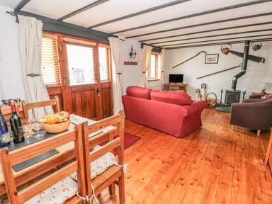 A living room with wooden floor and beams with a red sofa two brown chairs a dining table set with fruit and wine bottles a wood stove and decor items on the wall at Cowslip Cottage in Rosemarket