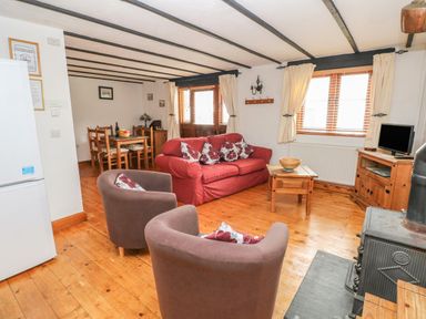 A living room with wooden floor a red sofa two brown chairs a wooden coffee table dining table and a wood burning stove at Cowslip Cottage in Rosemarket