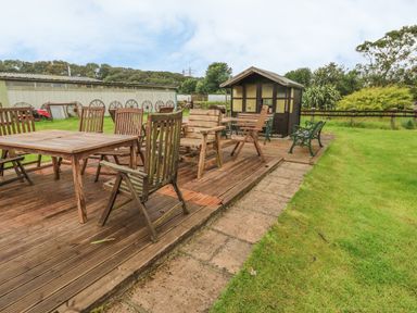 An outdoor wooden deck with wooden chairs and tables next to a small shed and green grass at Primose Cottage in Rosemarket