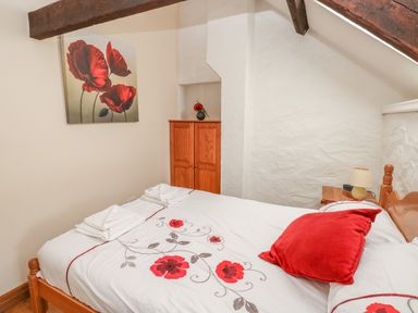 A bedroom with a bed with red flower bedding and a red pillow wooden wardrobe and a bedside table with a lamp at Primose Cottage in Rosemarket