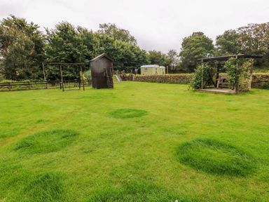 An outdoor grassy area with a wooden swing set a small dark wooden shed a play structure and a covered seating area with plants at Shepherds Hut - The Crook Rosemarket near Neyland