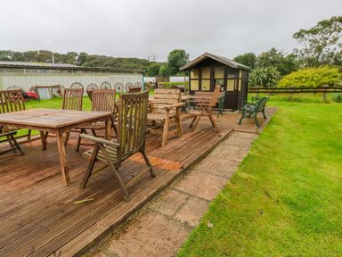 An outdoor wooden deck with wooden tables and chairs next to a small wooden cabin and a green lawn at Shepherds Hut - The Crook Rosemarket near Neyland