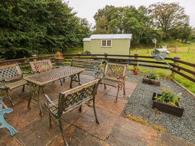 An outdoor patio with metal benches and table near a fenced area with a green shepherds hut at Shepherds Hut - The Crook Rosemarket near Neyland